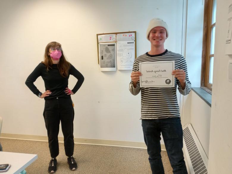 a student in a white hat holds a certificate while a woman looks on